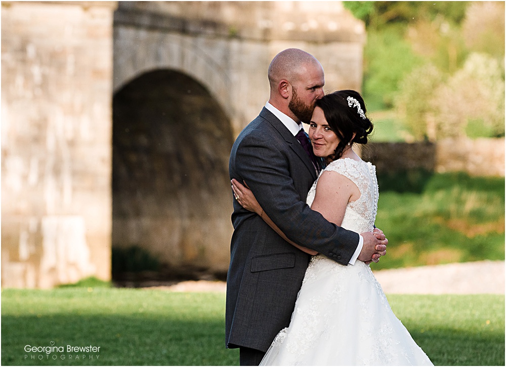 Bride and groom embrace near stone bridge on wedding day