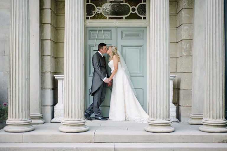 Bride and groom kissing by elegant pillar entranceway.