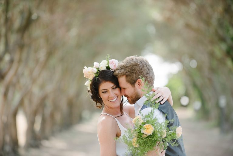 Bride and groom embrace under tree-lined path.