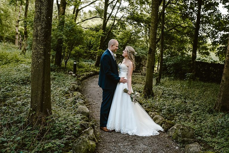 Bride and groom embrace in scenic woodland pathway.