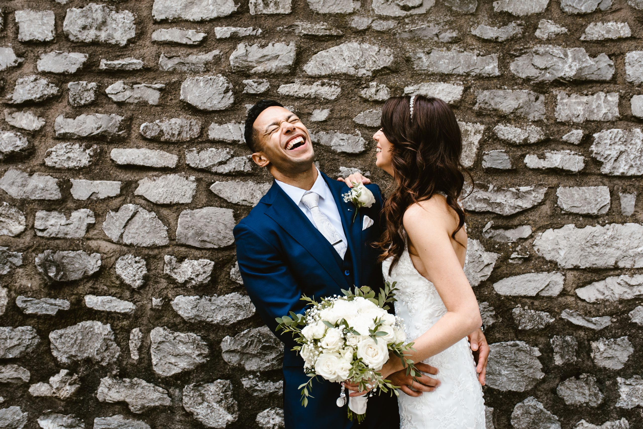 Bride and groom laughing joyfully against stone wall backdrop