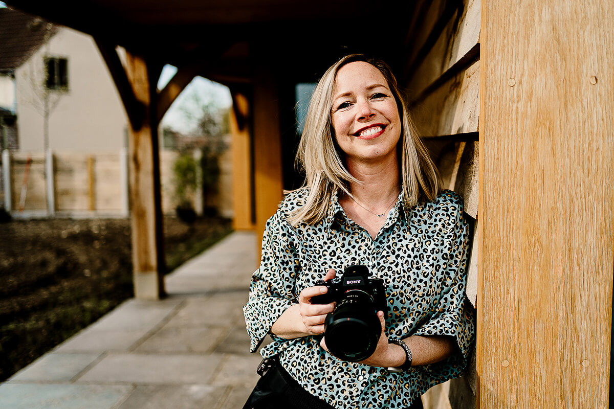 Woman smiling with camera leaning against wooden wall