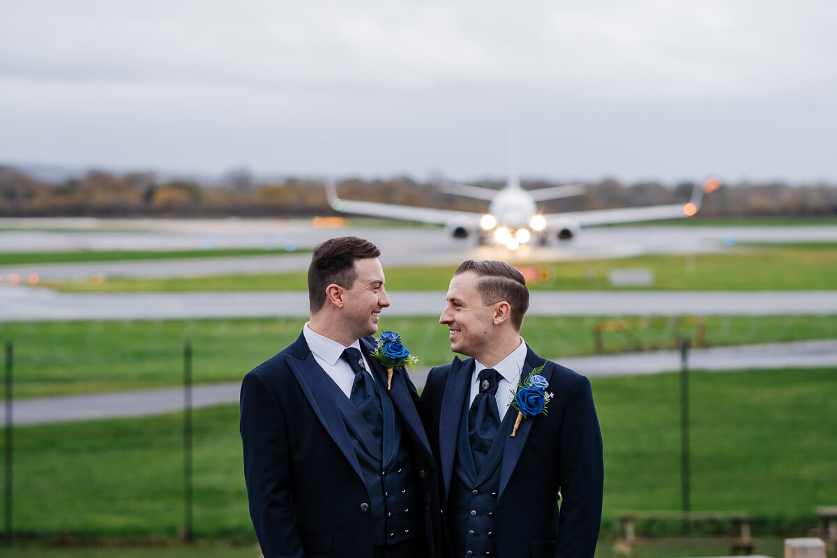 Two grooms smiling with a plane taking off in the background